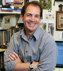 Waist-high image of John Bodinger de Uriarte sitting in his office. Books on shelves in the background.