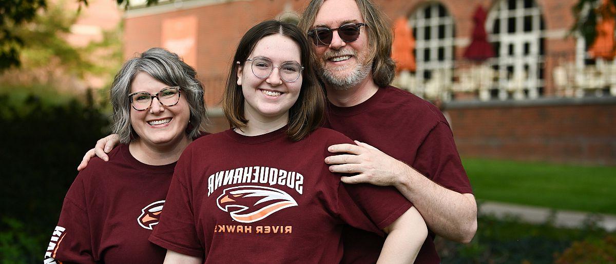 Image of mother, father and daughter posing and smiling at the camera. Brick building in the background.